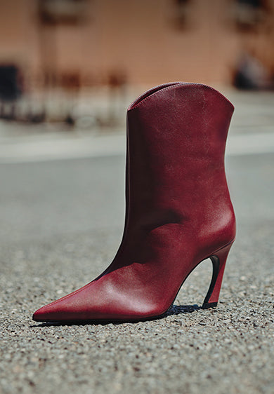 "A sleek, pointed-toe red leather ankle boot with a high heel standing upright on an empty asphalt road in an urban environment, with 'RED STYLES' text on the left side.