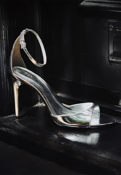 A shiny metallic silver high-heeled sandal with a delicate ankle strap is placed elegantly against a glossy black surface, reflecting light dramatically. Part of the "Metallics" selection.