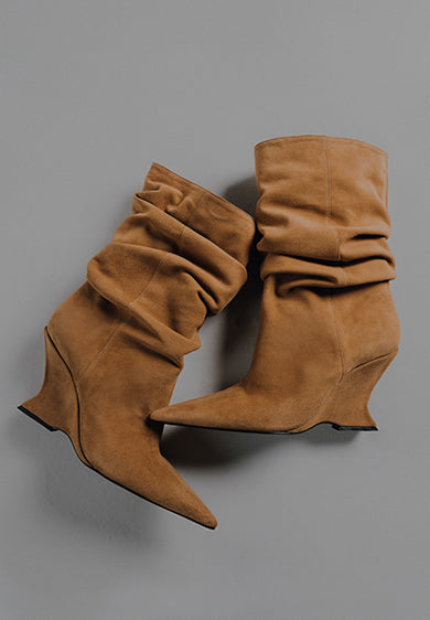 An overhead shot of a pair of slouchy, pointed-toe, tan suede ankle boots (booties) featuring a curved or sculptural wedge heel. The text "BOOTS & BOOTIES" is overlaid on the left side of the gray background.