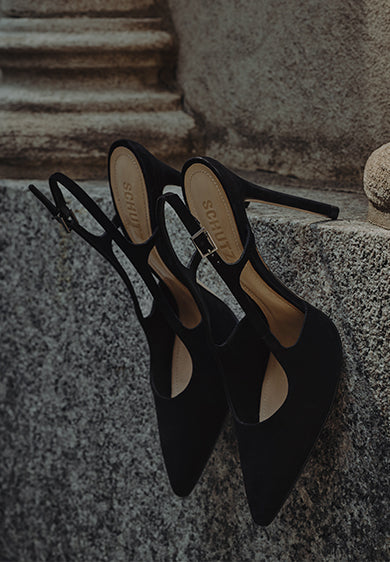 Black pointed-toe slingback heels resting on a stone ledge, with thin straps and a minimalist, elegant design.
