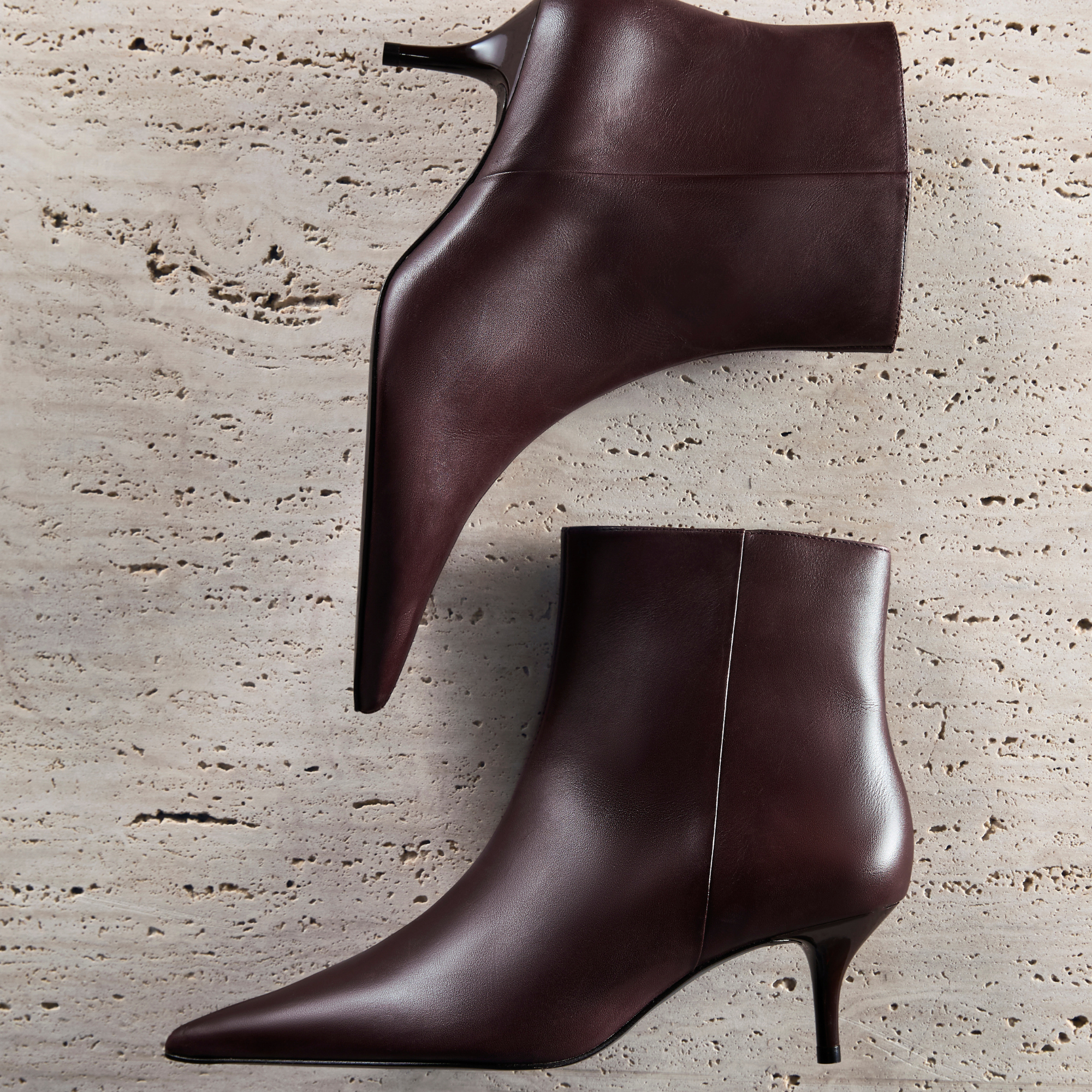 BEST SELLERS: Image shows two maroon leather ankle boots with pointed toes and kitten heels on a textured travertine-like stone background. A "SHOP NOW" link is visible below the main text.