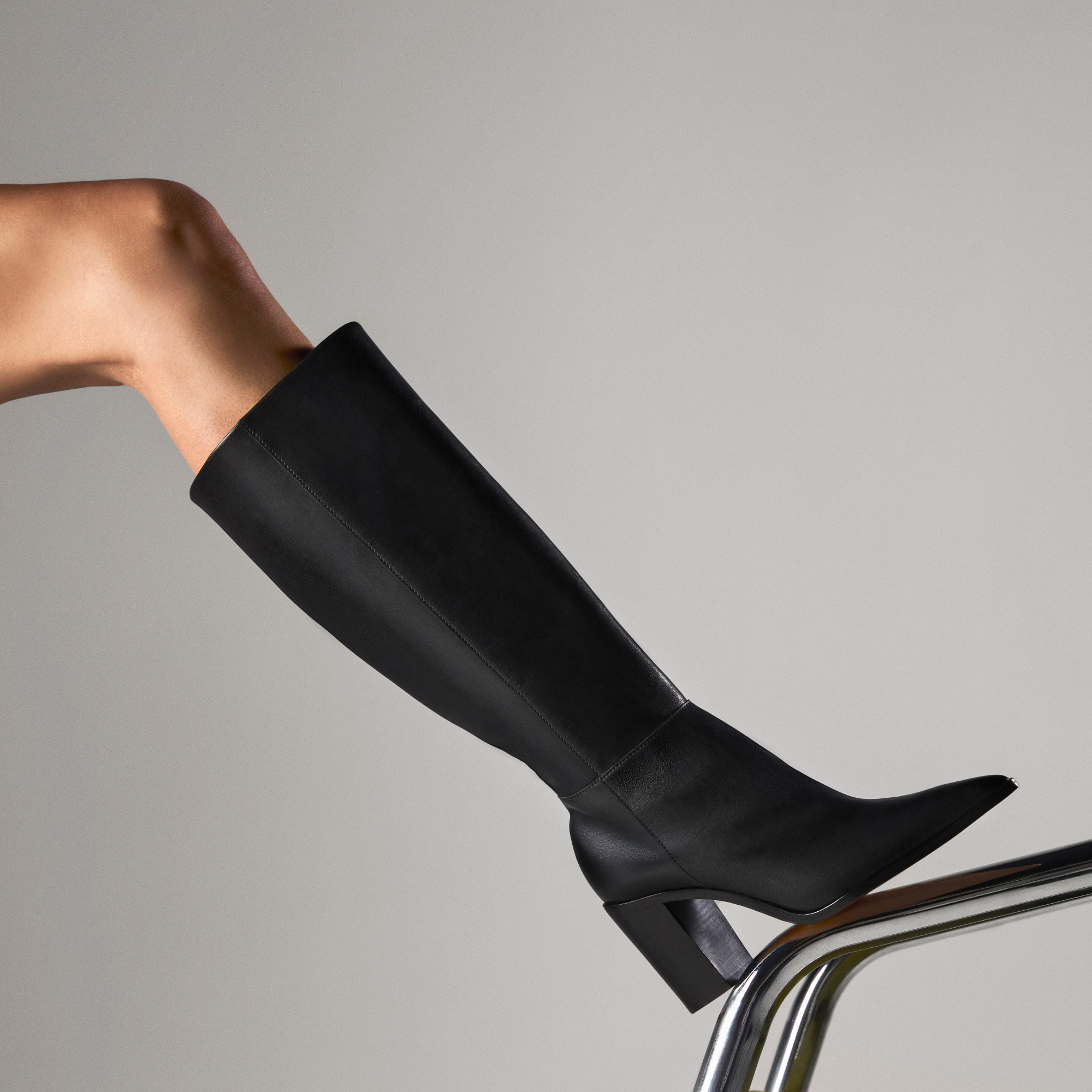 ESSENTIALS: Close-up image of a person wearing a black knee-high leather boot with a pointed toe and a flared block heel, leaning on a chrome rail. Includes a "SHOP NOW" link.