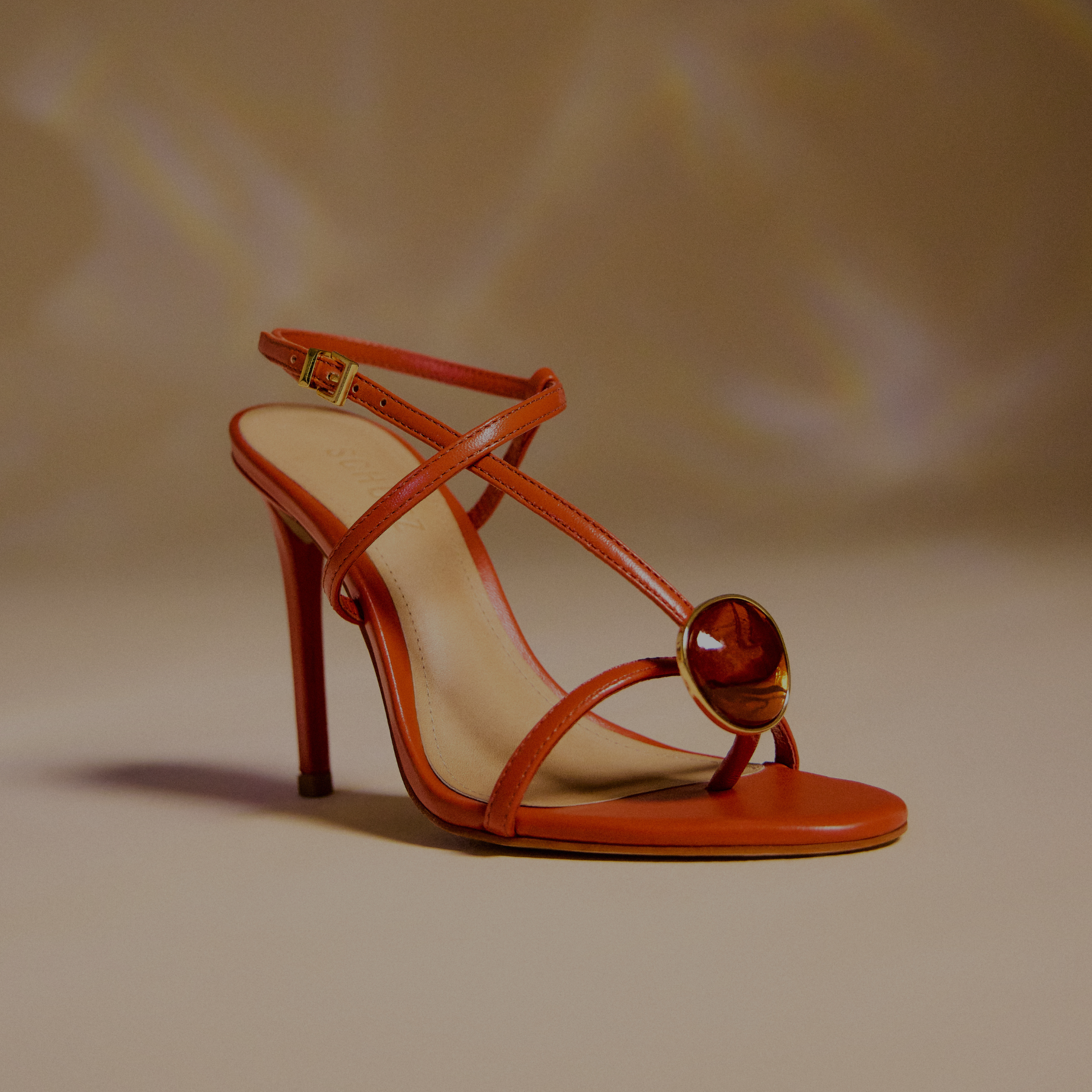 Black high-heel sandals with a sleek minimalist design, featuring thin straps and a stiletto heel. The pair is positioned on a reflective teal surface with soft indoor lighting highlighting the glossy finish.