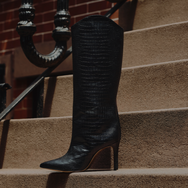 Two tall boots arranged side by side against a neutral wall. One boot is crafted in rich brown leather, and the other in glossy black leather. Both have pointed toes and high heels, shown under angled lighting that emphasizes their texture.