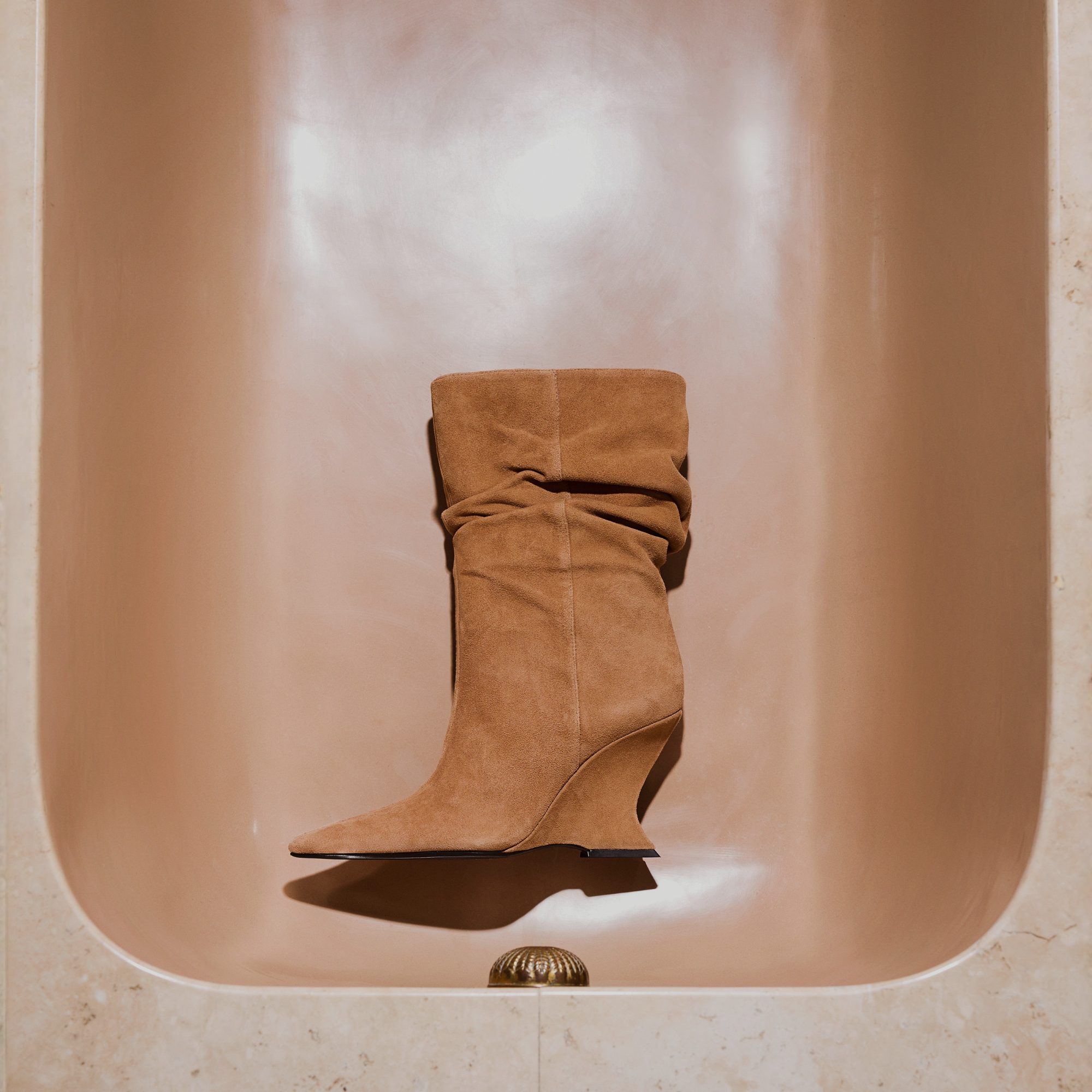 Advertisement for winter footwear featuring a single tan suede slouch boot with a wedge heel placed inside a beige bathtub. Text overlay: "WINTER ESCAPES" and a "SHOP NOW" button.
