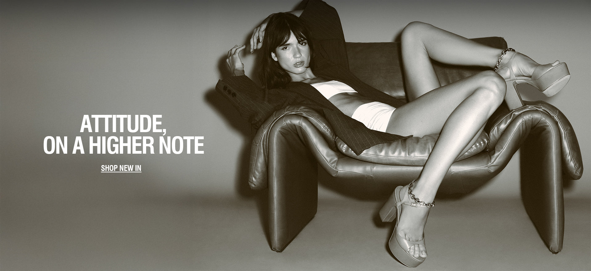 Model reclining on a leather armchair wearing a white bodysuit, tailored blazer, and platform heels, with the text “Attitude, on a higher note” and “Shop new in” displayed.