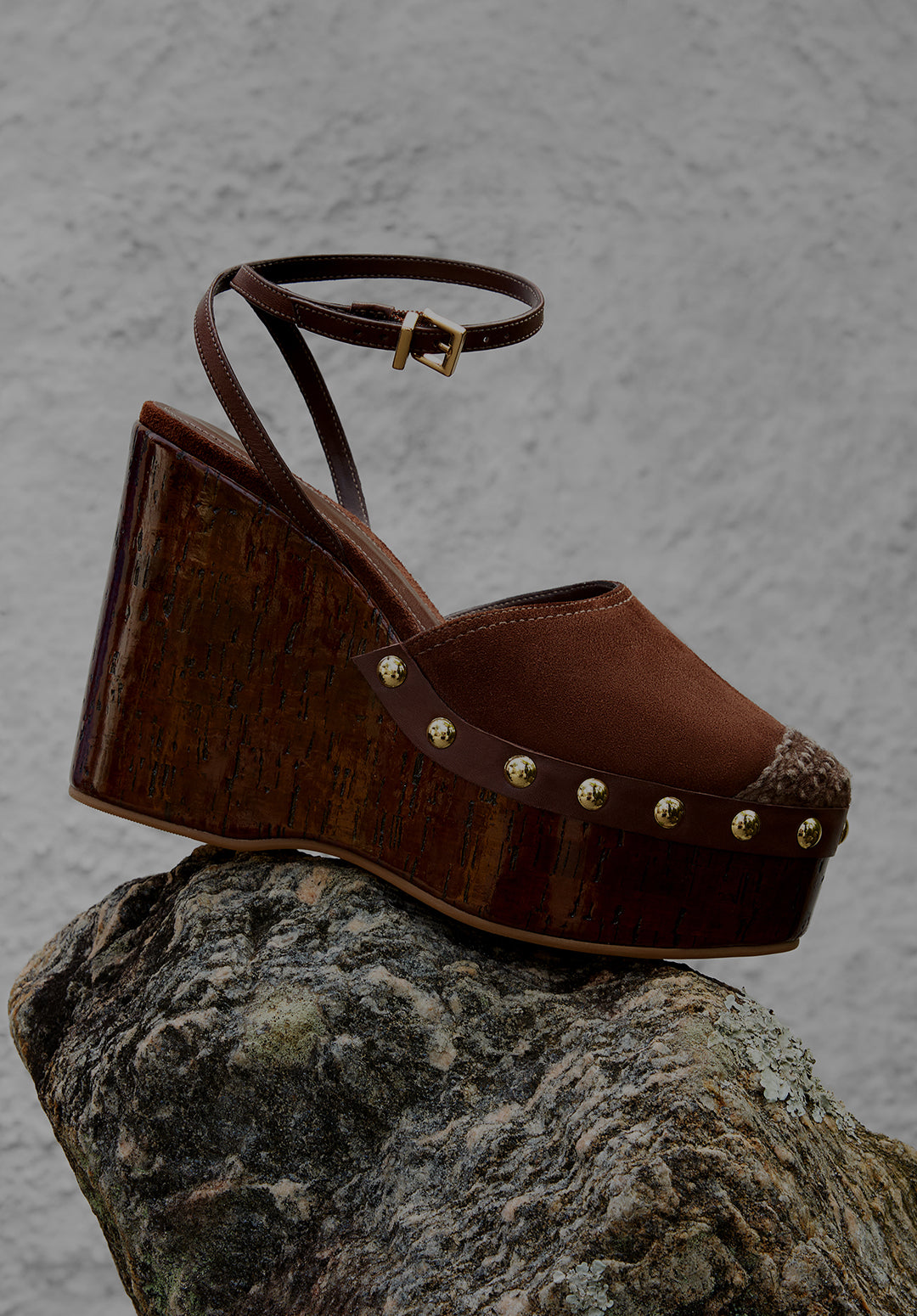
Brown platform wedge sandal with stud details and ankle strap displayed on a rock surface, highlighting bold platform and wedge styles.