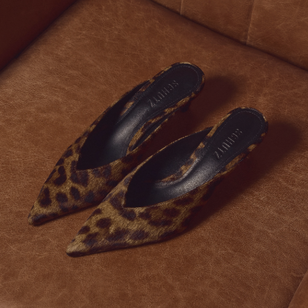 A pair of black leather pointed-toe mule heels with mid-height stiletto heels, displayed on a textured beige suitcase against a vintage brown suitcase background.