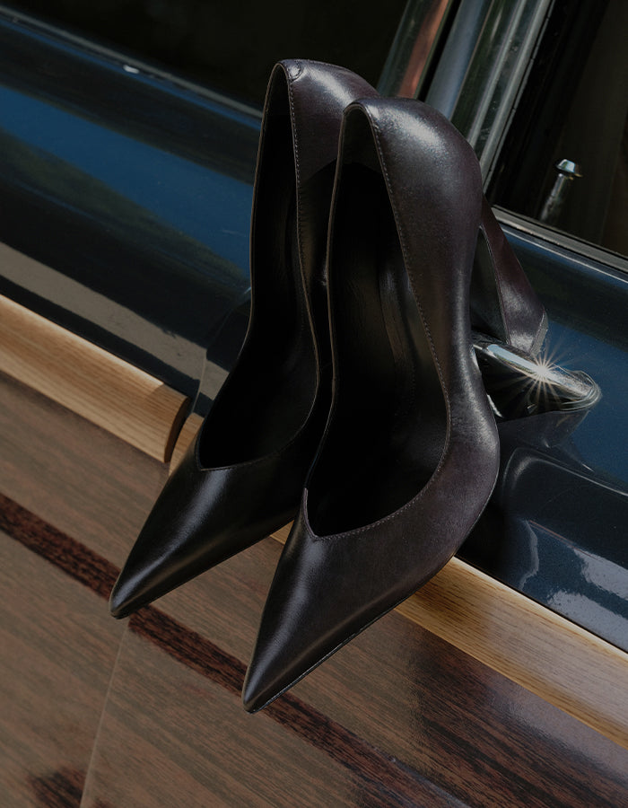 A pair of dark brown leather pointed-toe pumps with mid-height stiletto heels, resting on a polished wooden surface next to a dark car door.