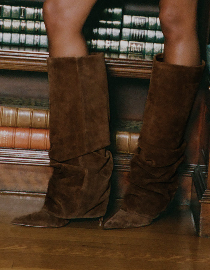 A person wearing tall brown suede slouch boots with pointed toes, standing on a wooden floor in front of a bookshelf.