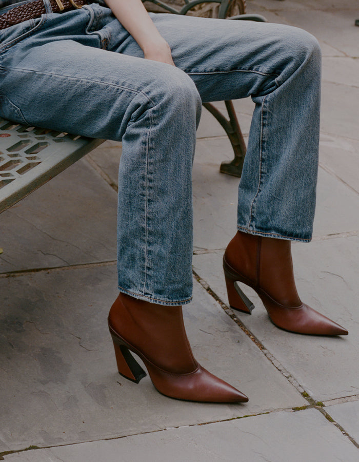 A person seated on a bench wearing brown pointed-toe heeled ankle boots with dark block heels, paired with light blue jeans on a stone-paved surface.
