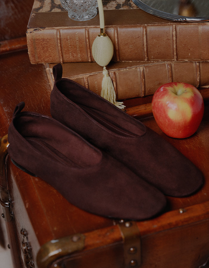 A pair of dark brown suede slip-on shoes displayed on a vintage wooden trunk, styled with old books, a tassel, and a red apple.